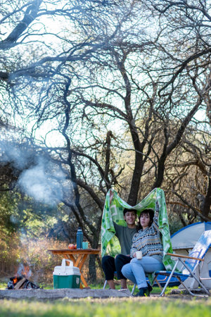 Two funny and cold friends drinking tea covered with a blanket. Campfire at the campsiteの写真素材