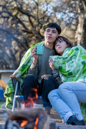 Two funny and cold friends drinking tea covered with a blanket. Campfire at the campsite