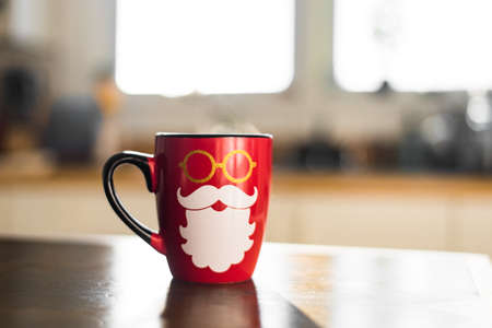Cute Festive Holiday Photo Of Bright Red Seasonal Santa Christmas Coffee Mug with Hipster Beard and Hot Steam Coming Off Cup in Light White Modern Kitchen with Sunshine Such a Warmの写真素材