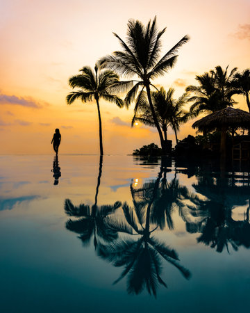 Silhouette of Beautiful Woman Walking on Edge of Infinity Pool in Vacation Paradise of Hawaii with Tropical Island Background of Palm Trees and Colorful Sunset Sky at Dusk with Refの写真素材