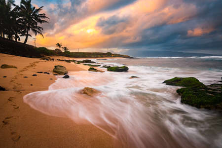 Tranquil Footprints in the Sand Long Exposure of Relaxing Tropical Island Paradise with Smooth Ocean Water Coming over Rocks on Sandy Beach with Colorful Pastel Sunrise Sky at Dawnの写真素材