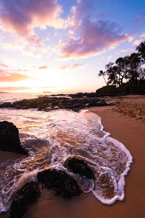 Tropical Island Paradise with Awesome Colorful Sky at Sunset and Ocean Water Coming over Rocks on Sandy Beach Shore with Palm Trees Silhouette at Secret Beach in Maui Hawaiiの写真素材