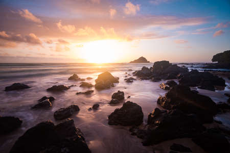 Relaxing Tropical Island Paradise of Colorful Pastel Sunrise Sky at Dawn with Sun Coming Over Ocean Horizon on Sandy Beach with Big Rocks and Mountain Background and Pink Cloudsの写真素材