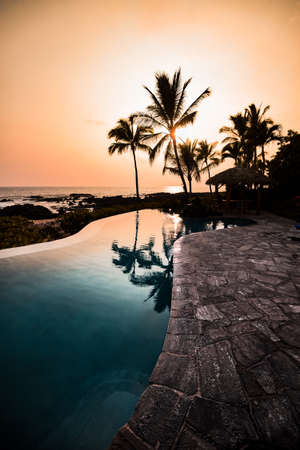 Aqua Blue Infinity Pool of a Vacation Destination Looking Out at Perfect Tropical Island Paradise of Palm Trees Silhouettes and Awesome Colorful Sunset Sky Over Ocean in Hawaiiのeditorial素材