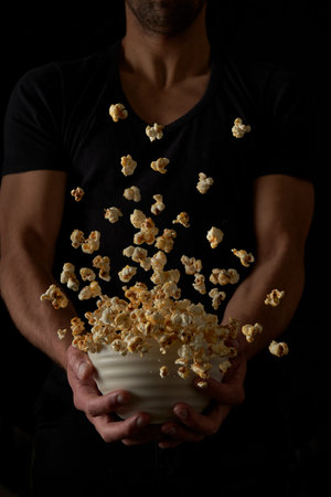 Unrecognizable man hold a white bowl of caramel pop corn flying and falling on isolated over black backgroundの写真素材