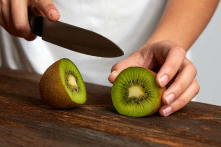 Unrecognizable woman cutting fresh ripe kiwi in half on wooden board.Front viewの写真素材