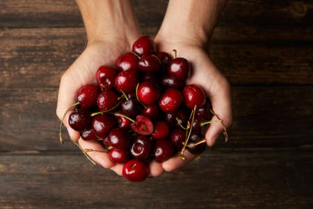 Unrecognizable man showing with his hands ripe cherries on wooden background. Top view. Food and...
