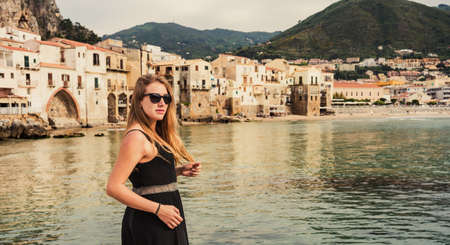 Beautiful young blonde woman with sunglasses in a photo session at the beach of the medieval village of Sicily island, Province of Palermo, Italy on a cloudy spring day.の写真素材