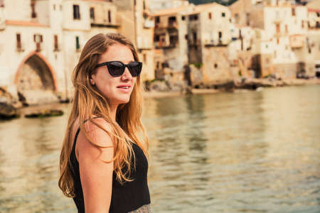 Beautiful young blonde woman with sunglasses smiling at the beach of the medieval village of Sicily island, Province of Palermo, Italy on a cloudy spring day.の写真素材
