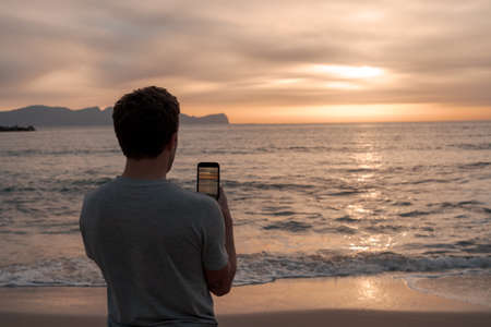 Young man on his back on vacation taking a picture with his phone of the amazing sunset in sicily, Italy. Travel conceptの写真素材