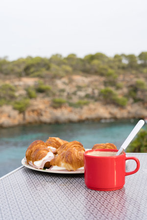 Breakfast of Coffee and croissant with ham and cheese in the outdoors with the sea in the background.の写真素材