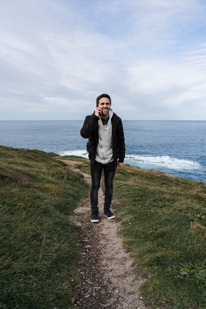 Smiling man talking on mobile phone while standing outdoors. sea scenery in the background. High quality photoの写真素材