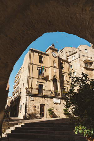 Historic building with clock tower in Gangi, Sicily viewed through a stone arch