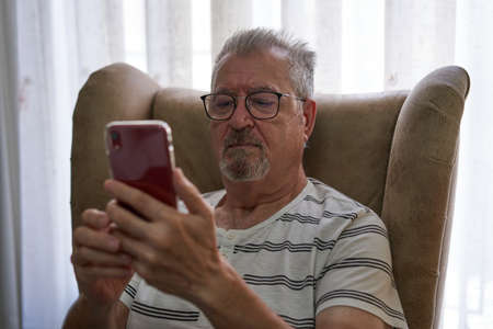 Older man is sitting in an armchair using his smartphone. He wears glasses and a beard and a striped...