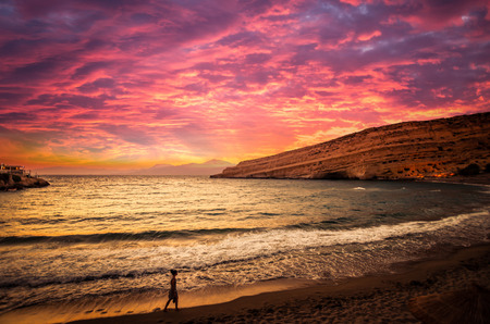 Sunset at Matala beach on Crete island, Greece. There is a girl walking on the beach. The colors in the sky are very beautiful, yellow, orange and red.の写真素材