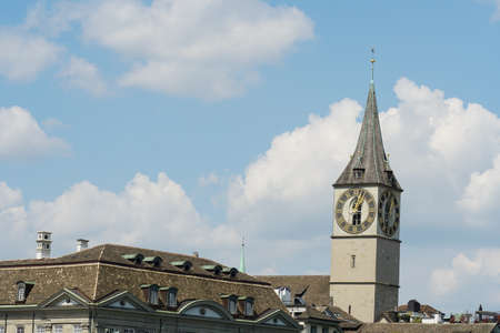 The tower of St. Peters Church in Zurich, Switzerland with its big clock and the rooftops of the surrounding buildings. On the background there is blue sky with white romantic -looking cumulus clouds.のeditorial素材
