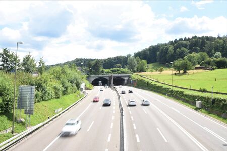 Motor highway with running cars and tunnel Honeret in Switzerland between Dietikon and village Urdorf. Express road, autobahn is surrounded by green slopes during summer day with blue sky and white cloudsの写真素材