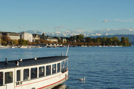 Lake Zurich in early October with a boat and the silhouette of the city on the left bank with small boats anchored on the foreground and the Alps with snow covered peaks on the background.のeditorial素材