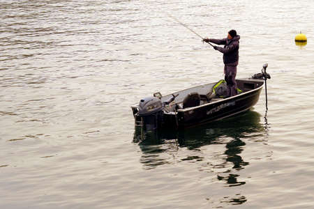 Zurich, Switzerland, - 02 06 2021: A hobby fisherman casting the fishing rod on the lake Zurich in winter. He is standing in a boat and there is a yellow buoy on the water surface around him.のeditorial素材