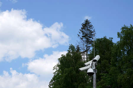 Cctv camera attached on a metal pole with coniferous trees and sky on the background and a lot of...