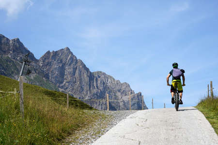 Man on a bicycle on a hilltop in rear view. There are Alp mountains on the left hand side. Cycling and hiking trail in the Engelberg region in Switzerland.の写真素材