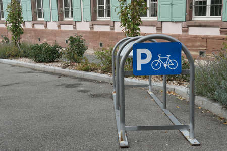 Arch metal frame for parking bicycles with blue sign containing letter P and pictogram of bicykle situated on the street near green house and pavement with vegetation of plants, grass and bushes.の写真素材