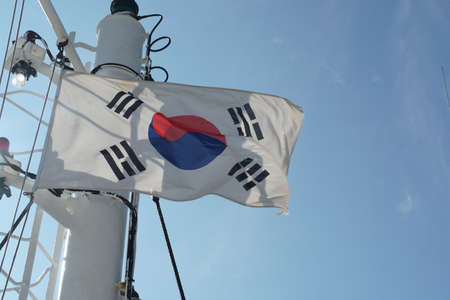 South Korean flag flapping on white mast of a merchant container ship with red and  white navigational lights situated in monkey radar island  during clear blue sky in summer day with copy space.の写真素材