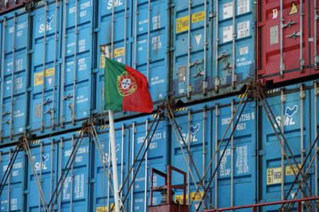 Flag of Portugal flapping on white mast situated in aft or stern part of container ship. There are stowed and lashed containers with bars and twist locks on deck in background.の写真素材