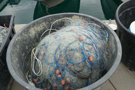 Fishing net with blue ropes and red-brown floats stored in black oval plastic bucket ready for...