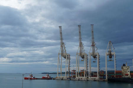 Koper, Slovenia - 05 07 2022: New modern very high gantry cranes in vertical position waiting for cargo operation under dramatic sky with dark blue clouds. On the right side is superstructure of ship.のeditorial素材