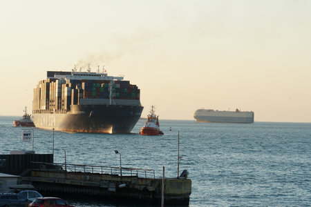 Koper, Slovenia - 04 16 2022: Huge fully loaded container ship from CMA CGM company entering port of Koper during sunset. Ship is assisted by orange tugboat through calm sea. In hoのeditorial素材