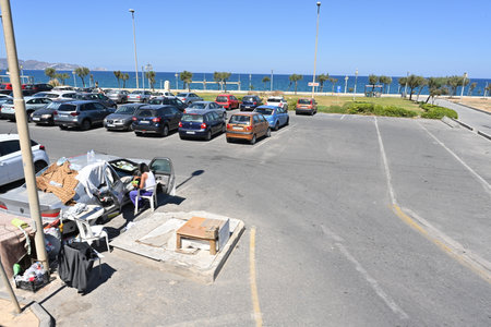 Parking lot on the coast full of cars. A man sitting on a plastic chair by his car and eats from a bowl.  He lives in the car. It is covered by pieces of textile.のeditorial素材