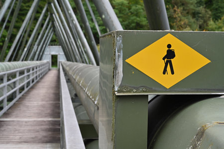 Yellow tourist sing with pictogram of  hiker on the timber work bridge crossing the river. To the bridge from both sides are attached two, green painted, metal pipelines.の写真素材