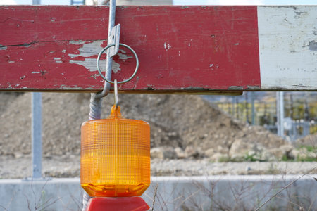 View on wireless warning construction light of beacon shape and orange color hanging from wooden red and white construction barrier. Behind is building ground with heap of earth.の写真素材