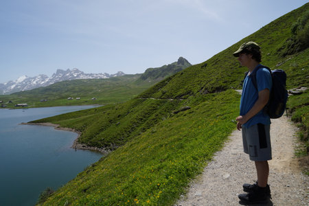 Teenage male hiker with a backpack is standing on a hiking trail over the lake. He is looking at the lake Melchsee enjoying the view. Behind are Swiss alps.のeditorial素材