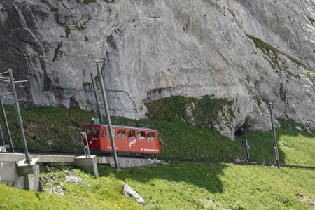 Red cogwheel train between railway  station Alpnachstad and mountain Pilatus Kulm. Behind are rocky cliffs of swiss alps. Track line are surrounded by green grass.の写真素材