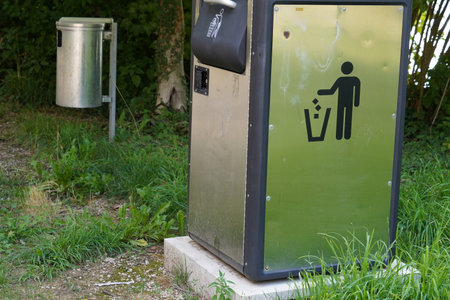 Two metal litter bins in public spaces. The first one is cuboidal and bears a icon instructing to put litter in it. The second one on the background is cylindrical.の写真素材