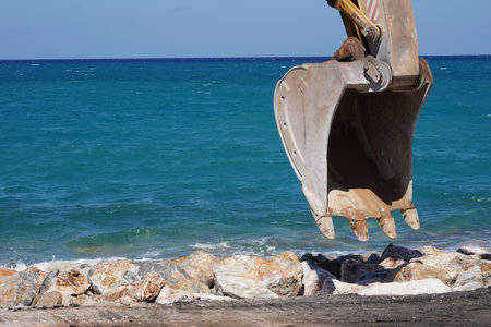 Close-up of a dipper bucket of  crawler excavator working on the construction site. Behind is a rock beach and Sea of Crete.の写真素材