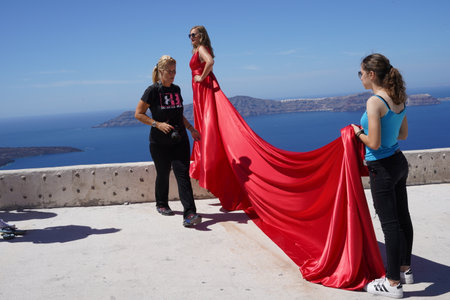 Santorin, Greece, 09 14 2023: An assistant are holding the model's train of a red dress. The model is standing on a wall and behind her is the sea and an island.のeditorial素材