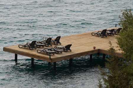 Wooden platform or pier sits by the seaside at the base of a steep incline, with vacant armchairs facing the sea.の写真素材