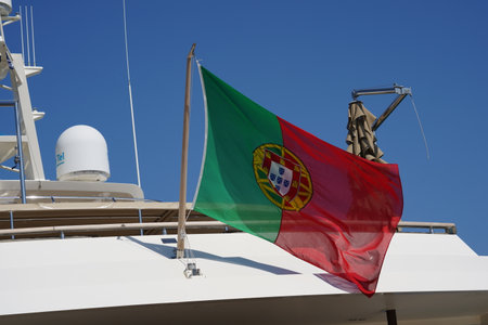 Portugal flag flapping on short wooden mast situated in aft or stern part of luxury white yacht. There are navigational and satelite antennas in background.の写真素材