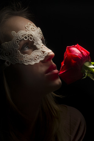 Portrait of a shy girl wearing mask and red rose on black backgroundの写真素材