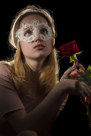 Portrait of a shy girl wearing mask and red rose on black backgroundの写真素材