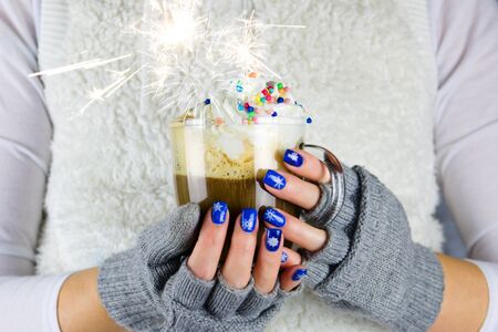 Woman with beautiful blue nails holding a mug of hot coffee and whipped cream with sugar decoration and sparkle. She has gray gloves without fingers.の写真素材