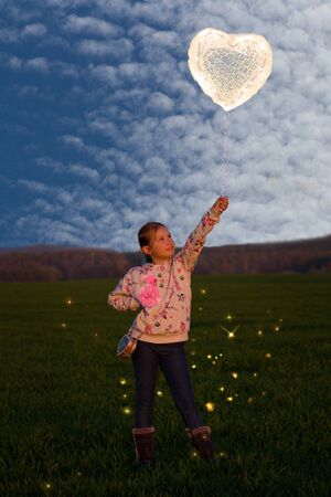 A little girl stands in the grass and holds a glowing heart-shaped balloon.の写真素材