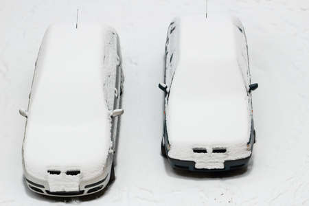 Two parked cars stand in a snowy parking lot and are covered with snow. A snowstorm struck. The cars and roads are under the snow.の写真素材