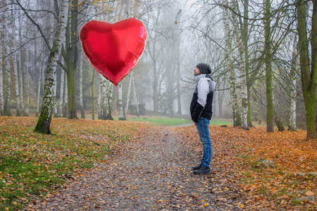 A young man stands in the middle of an autumn landscape and stares at a inflatable helium balloon heart. A red inflatable helium balloon hearts in a mysterious deciduous forest.の写真素材