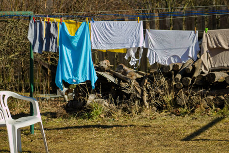 Laundry is drying on a clothesline in the countryside. A white plastic chair stands next to the clothesline. Washed laundry on a clothesline. T-shirts and socks are dried in the sun.の写真素材