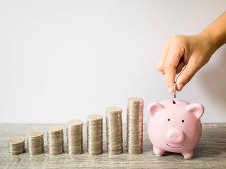 Woman hand putting coin into pink piggy bank with coins pile growth graph, saving money for future investment plan and retirement fund concept.の写真素材
