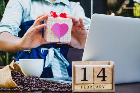 Woman hand holding gift box with laptop, 14 February wooden calendar, coffee cup and coffee beans. Valentine day concept.の写真素材